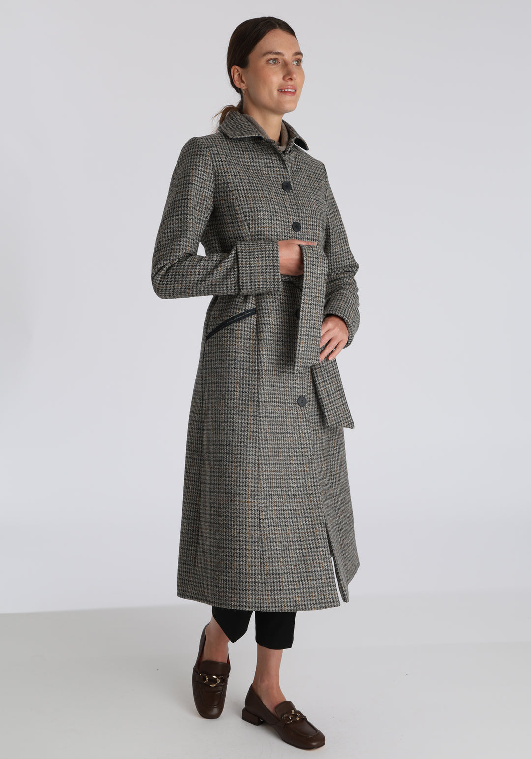 Model wearing the Belted coat in stone grey and ochre dogtooth check wool, a long tailored style with a matching belt, turn-back cuffs, and a deep back vent for ease of movement.