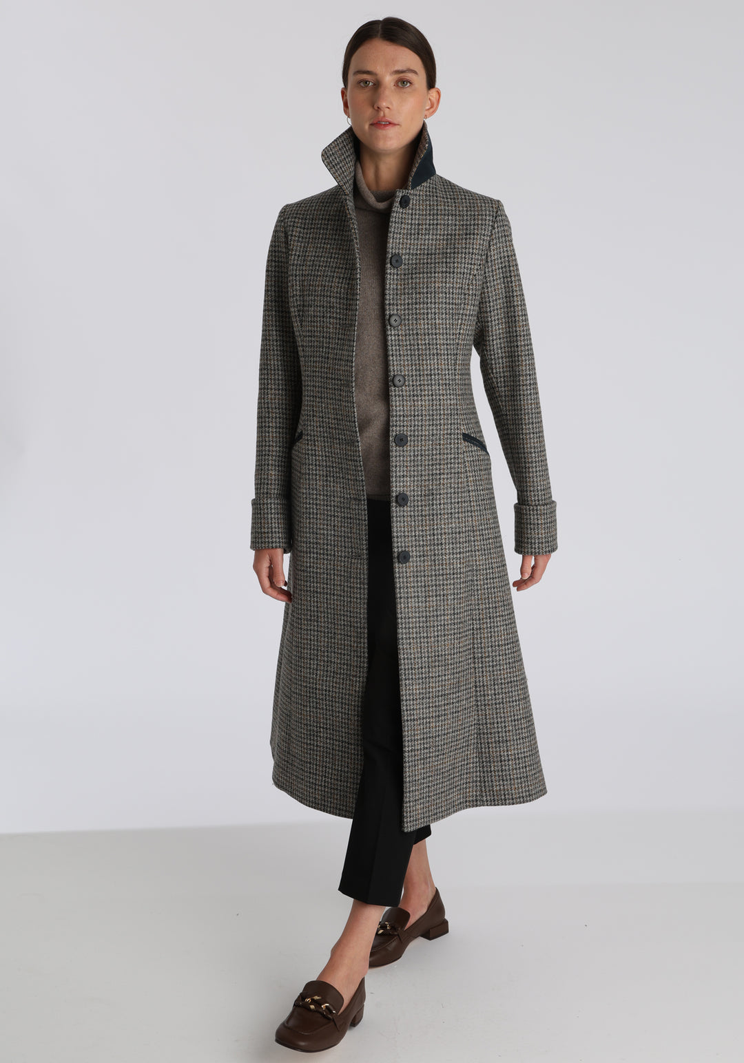 Model wearing the Belted coat in stone grey and ochre dogtooth check wool, a long tailored style with a matching belt, turn-back cuffs, and a deep back vent for ease of movement.