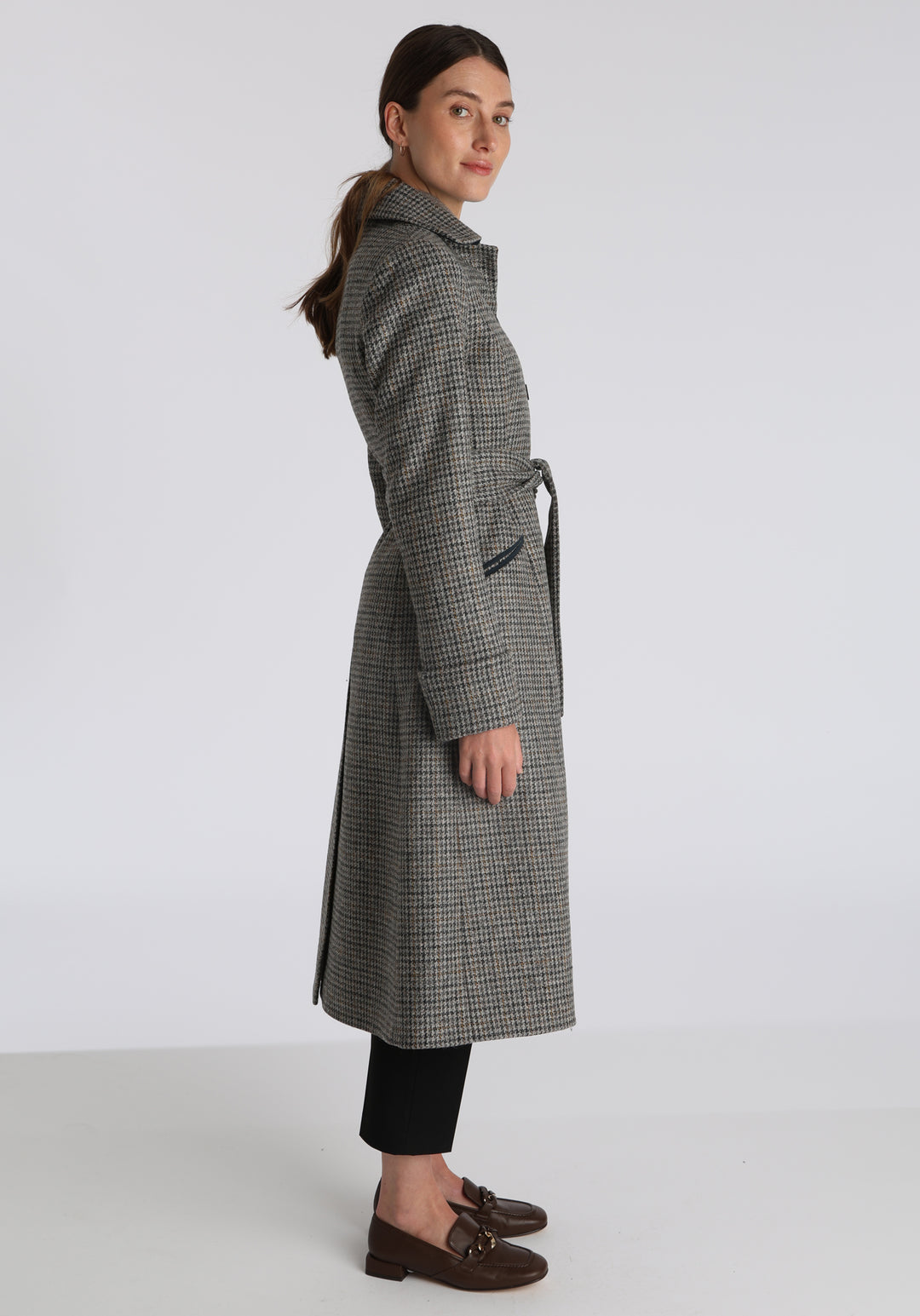 Model wearing the Belted coat in stone grey and ochre dogtooth check wool, a long tailored style with a matching belt, turn-back cuffs, and a deep back vent for ease of movement.