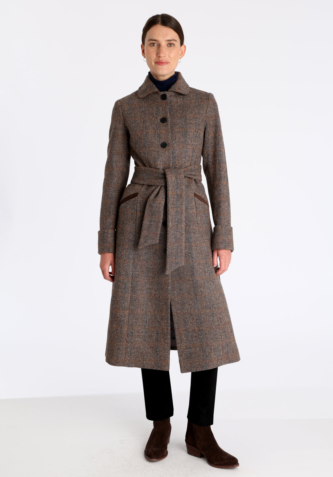 Model wearing the Belted coat in clay check Harris Tweed, a long tailored style with a matching belt, turn-back cuffs, and a deep back vent for ease of movement.