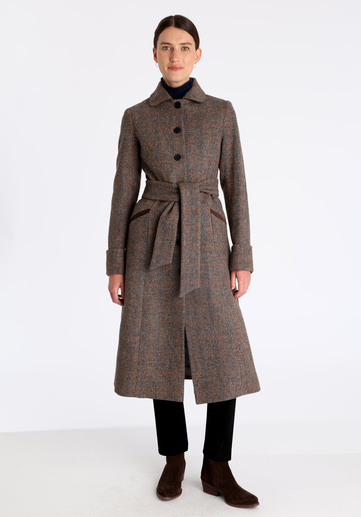 Model wearing the Belted coat in clay check Harris Tweed, a long tailored style with a matching belt, turn-back cuffs, and a deep back vent for ease of movement.