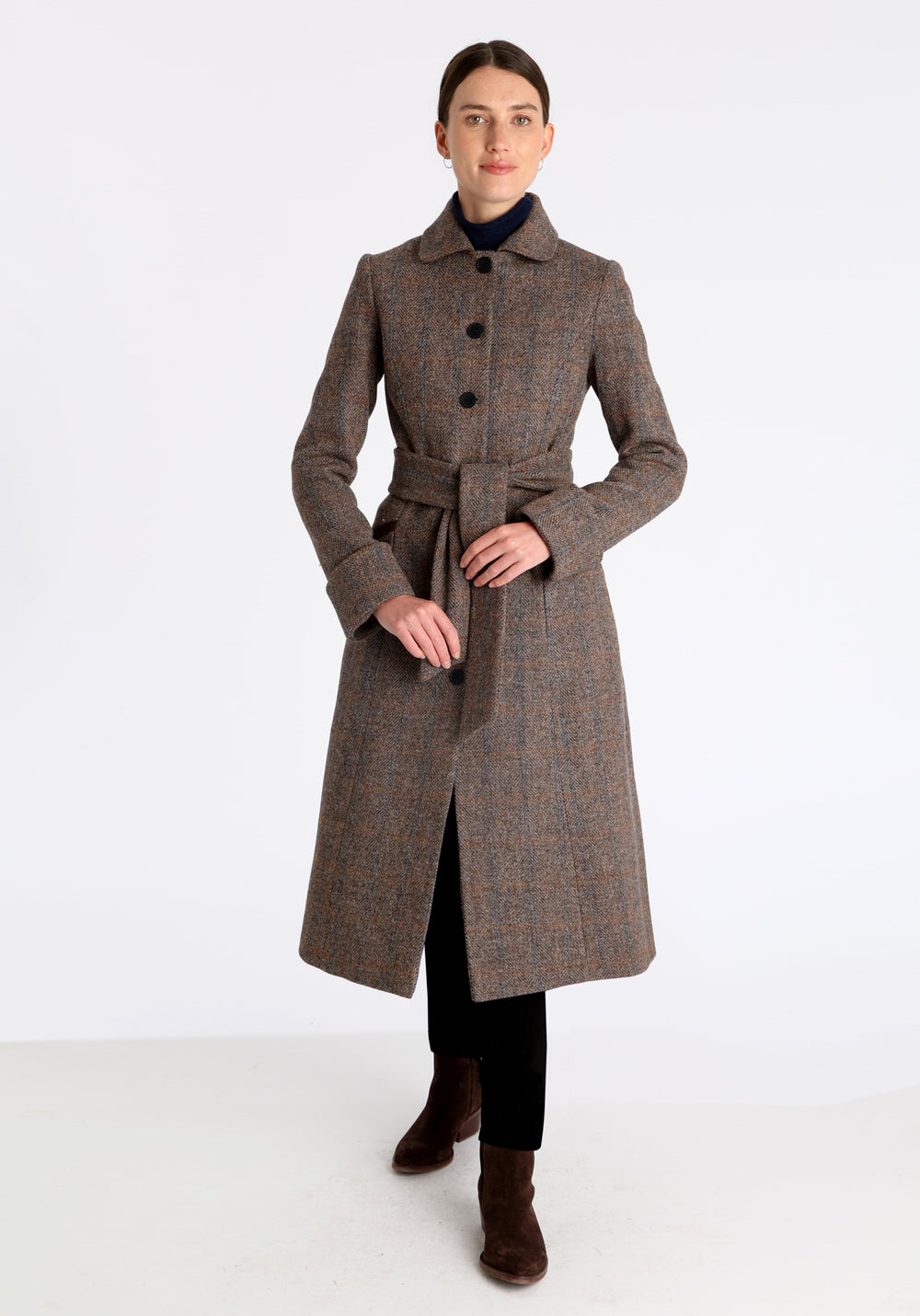 Model wearing the Belted coat in clay check Harris Tweed, a long tailored style with a matching belt, turn-back cuffs, and a deep back vent for ease of movement.