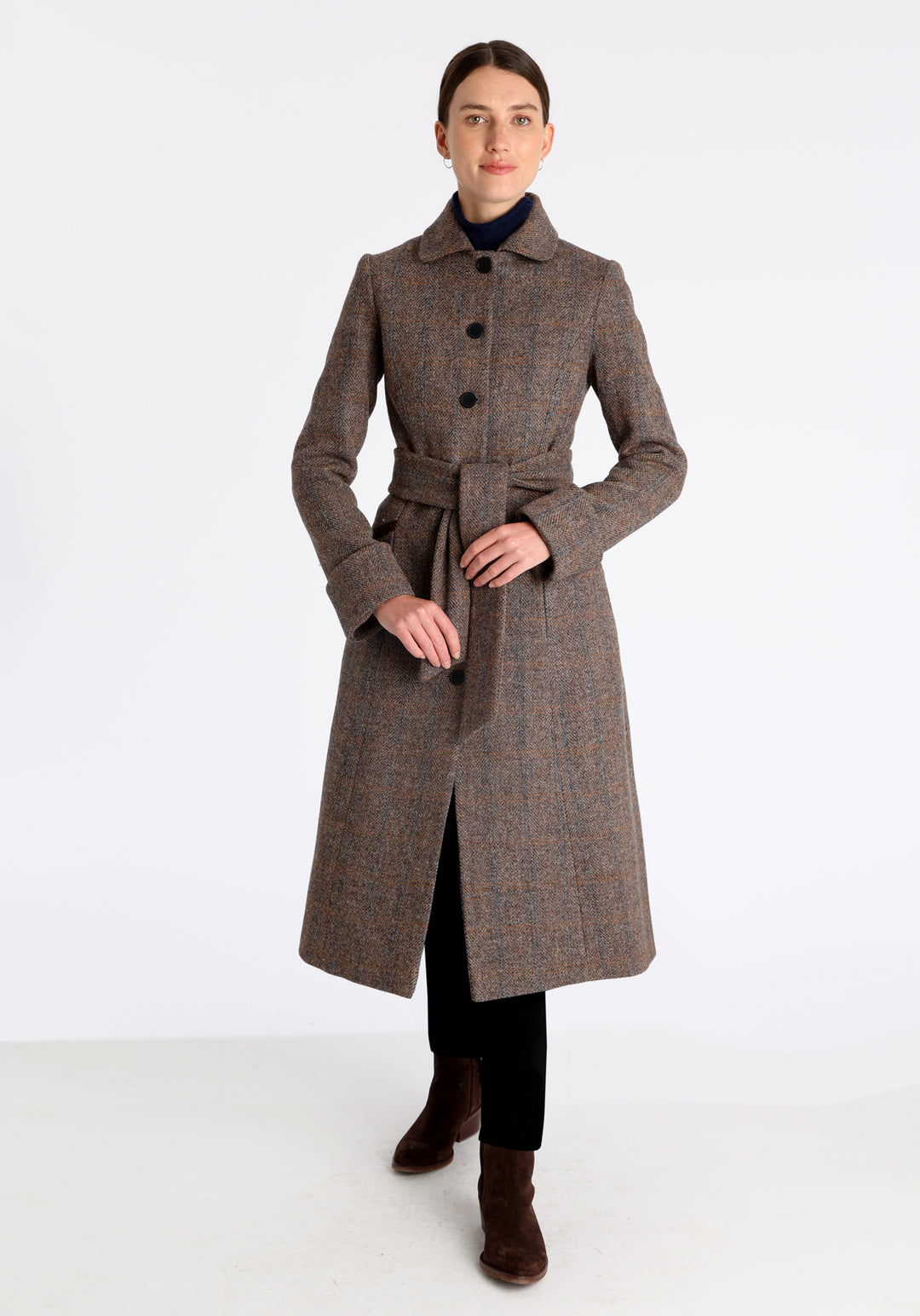 Model wearing the Belted coat in clay check Harris Tweed, a long tailored style with a matching belt, turn-back cuffs, and a deep back vent for ease of movement.