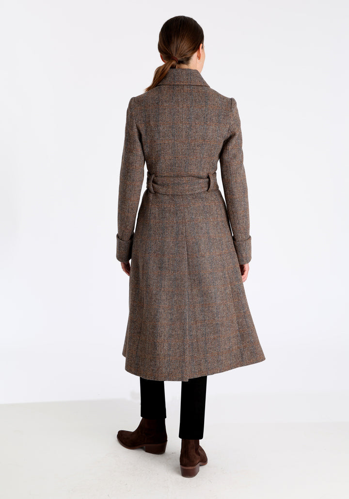 Model wearing the Belted coat in clay check Harris Tweed, a long tailored style with a matching belt, turn-back cuffs, and a deep back vent for ease of movement.