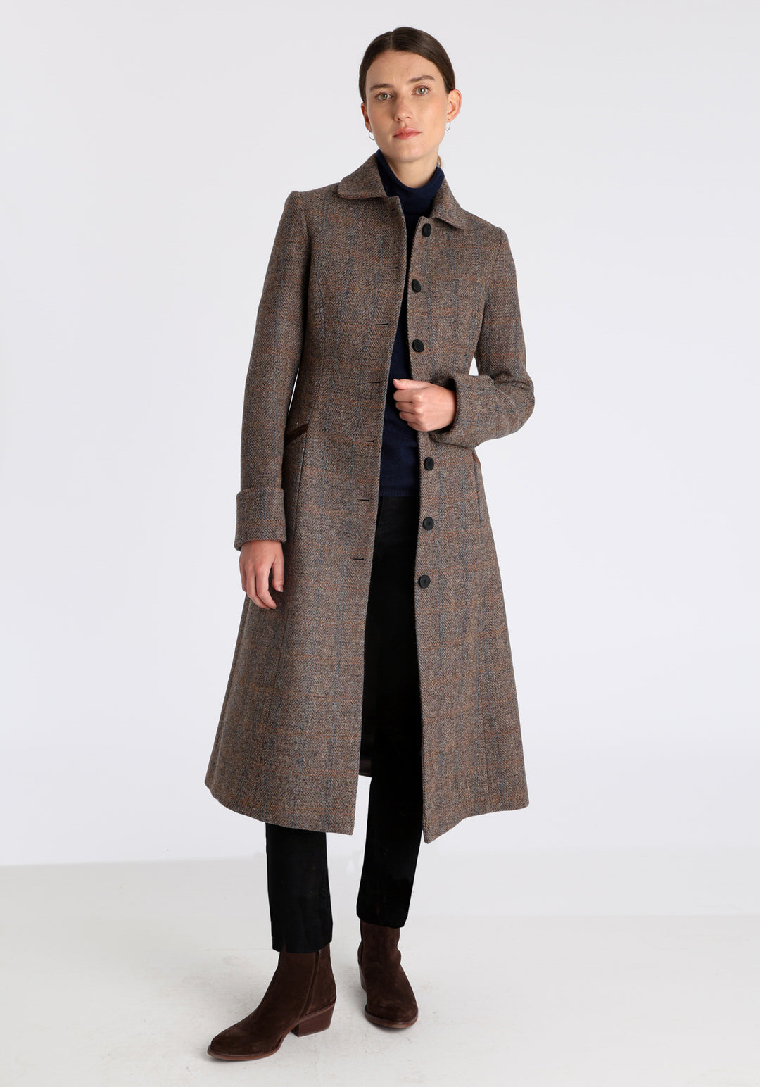 Model wearing the Belted coat in clay check Harris Tweed, a long tailored style with a matching belt, turn-back cuffs, and a deep back vent for ease of movement.