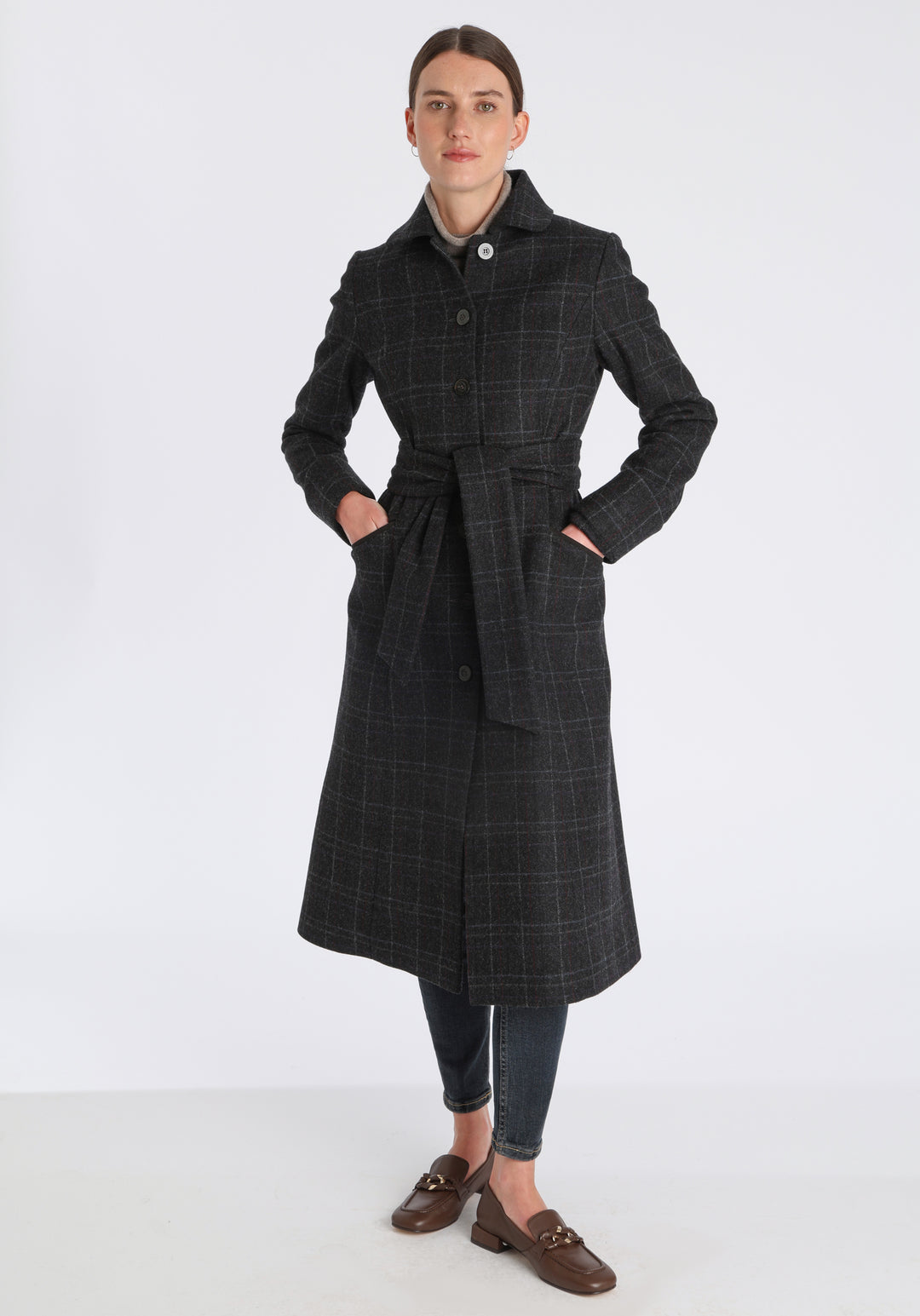 Model wearing the Belted coat in black with faint overcheck, a long tailored style with a matching belt, turn-back cuffs, and a deep back vent for ease of movement.