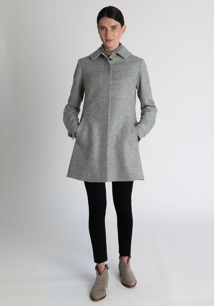 Model wearing the Cape coat in slate blue and cream twill lambswool, a short, structured style with a generous collar, buttoned cuffs, and a deep back pleat that creates a subtle cape like shape.