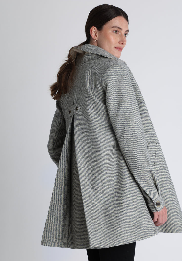 Model wearing the Cape coat in slate blue and cream twill lambswool, a short, structured style with a generous collar, buttoned cuffs, and a deep back pleat that creates a subtle cape like shape.