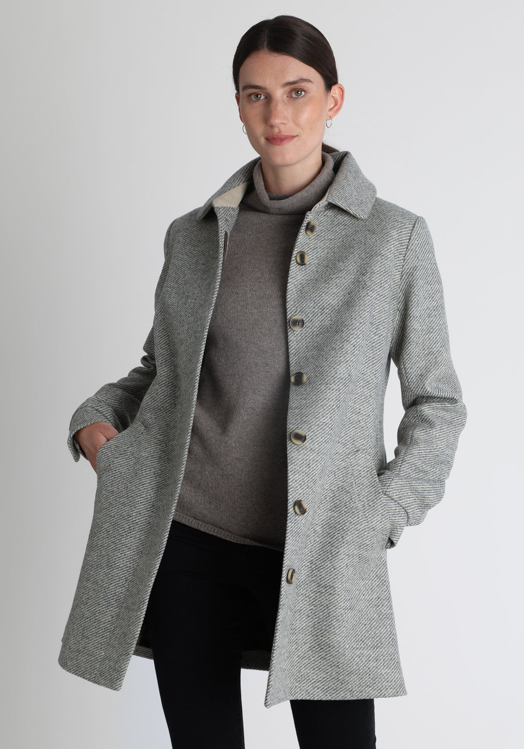 Model wearing the Cape coat in slate blue and cream twill lambswool, a short, structured style with a generous collar, buttoned cuffs, and a deep back pleat that creates a subtle cape like shape.