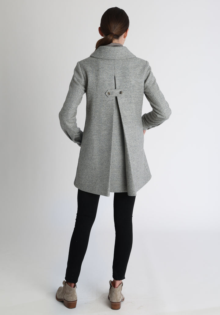 Model wearing the Cape coat in slate blue and cream twill lambswool, a short, structured style with a generous collar, buttoned cuffs, and a deep back pleat that creates a subtle cape like shape.