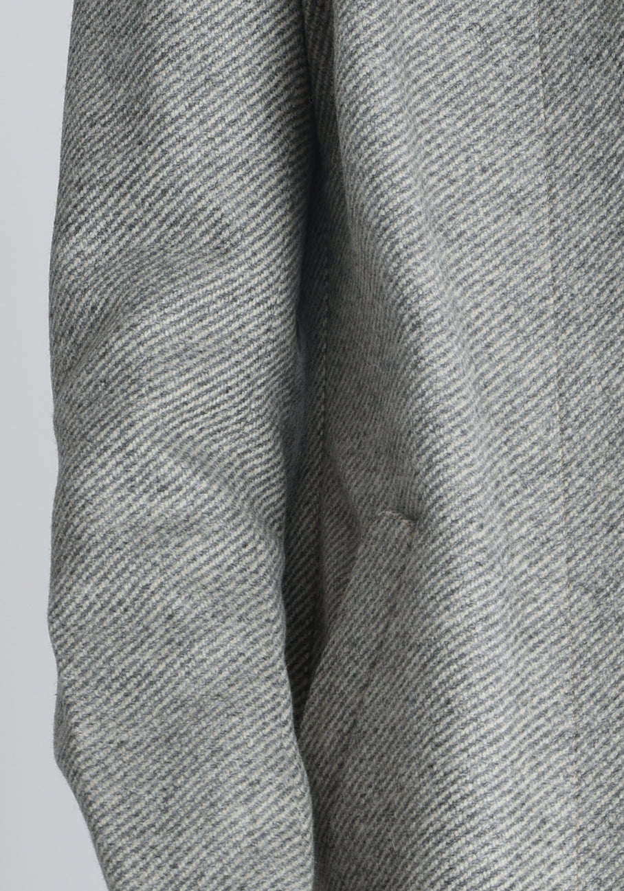 Model wearing the Cape coat in slate blue and cream twill lambswool, a short, structured style with a generous collar, buttoned cuffs, and a deep back pleat that creates a subtle cape like shape.
