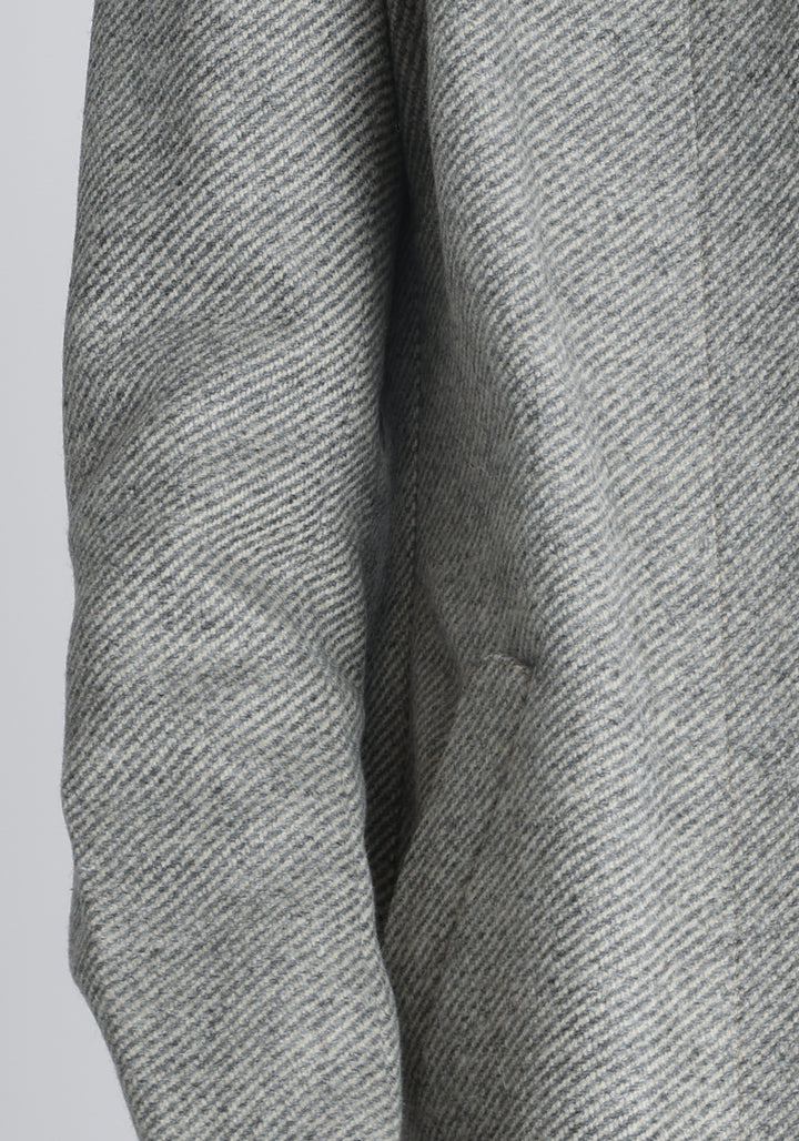 Model wearing the Cape coat in slate blue and cream twill lambswool, a short, structured style with a generous collar, buttoned cuffs, and a deep back pleat that creates a subtle cape like shape.