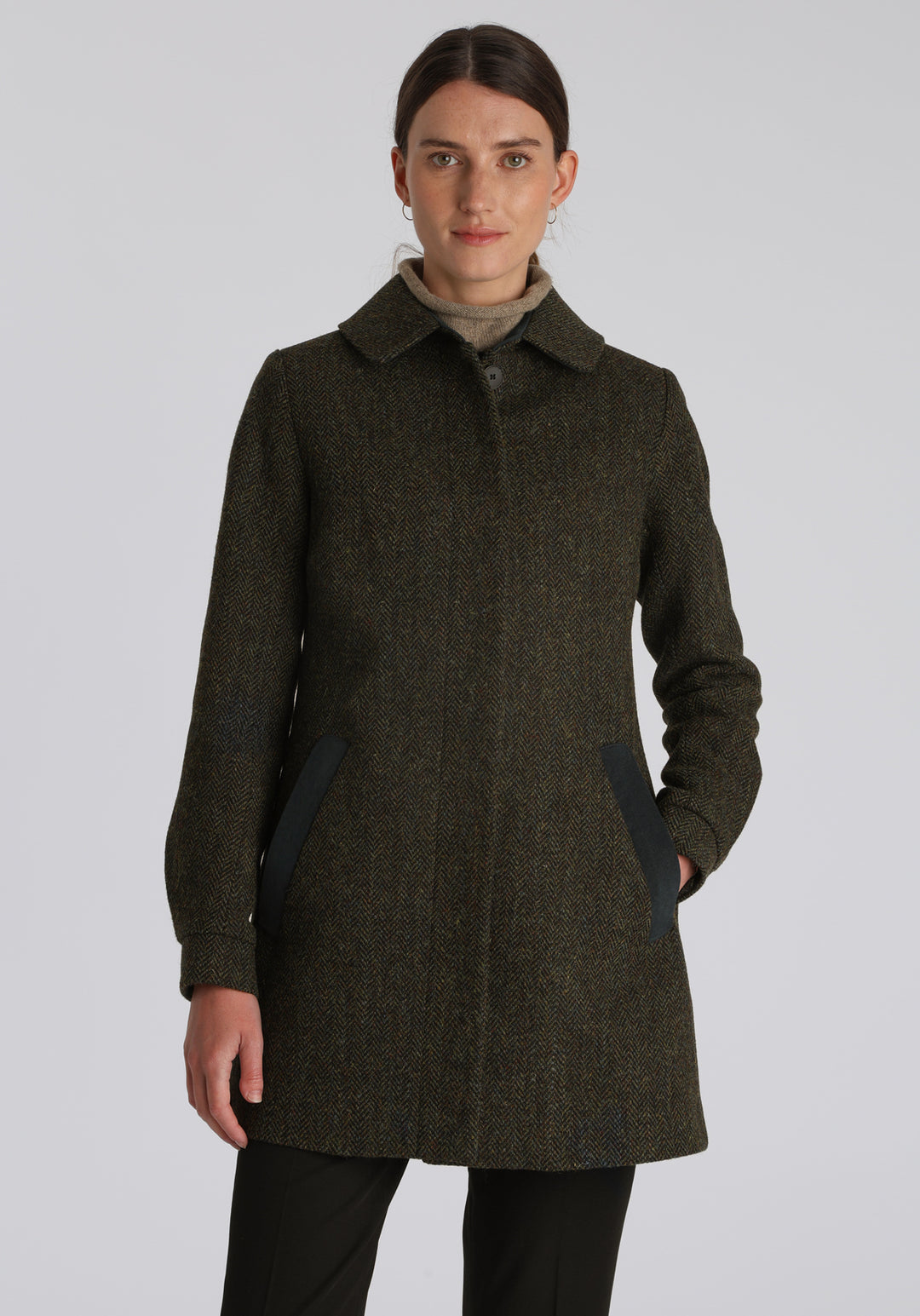 Model wearing the Cape coat in green spruce herringbone Harris Tweed, a short, structured style with a generous collar, buttoned cuffs, and a deep back pleat that creates a subtle cape-like shape.