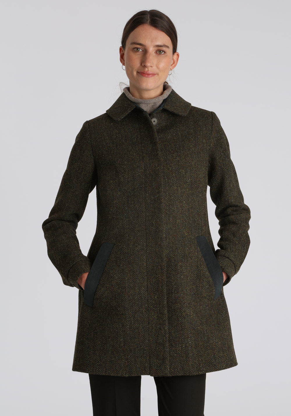 Model wearing the Cape coat in green spruce herringbone Harris Tweed, a short, structured style with a generous collar, buttoned cuffs, and a deep back pleat that creates a subtle cape-like shape.