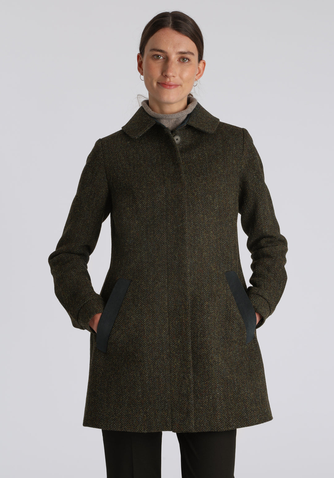 Model wearing the Cape coat in green spruce herringbone Harris Tweed, a short, structured style with a generous collar, buttoned cuffs, and a deep back pleat that creates a subtle cape-like shape.