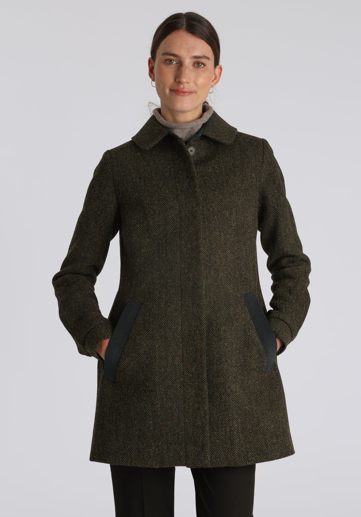 Model wearing the Cape coat in green spruce herringbone Harris Tweed, a short, structured style with a generous collar, buttoned cuffs, and a deep back pleat that creates a subtle cape-like shape.