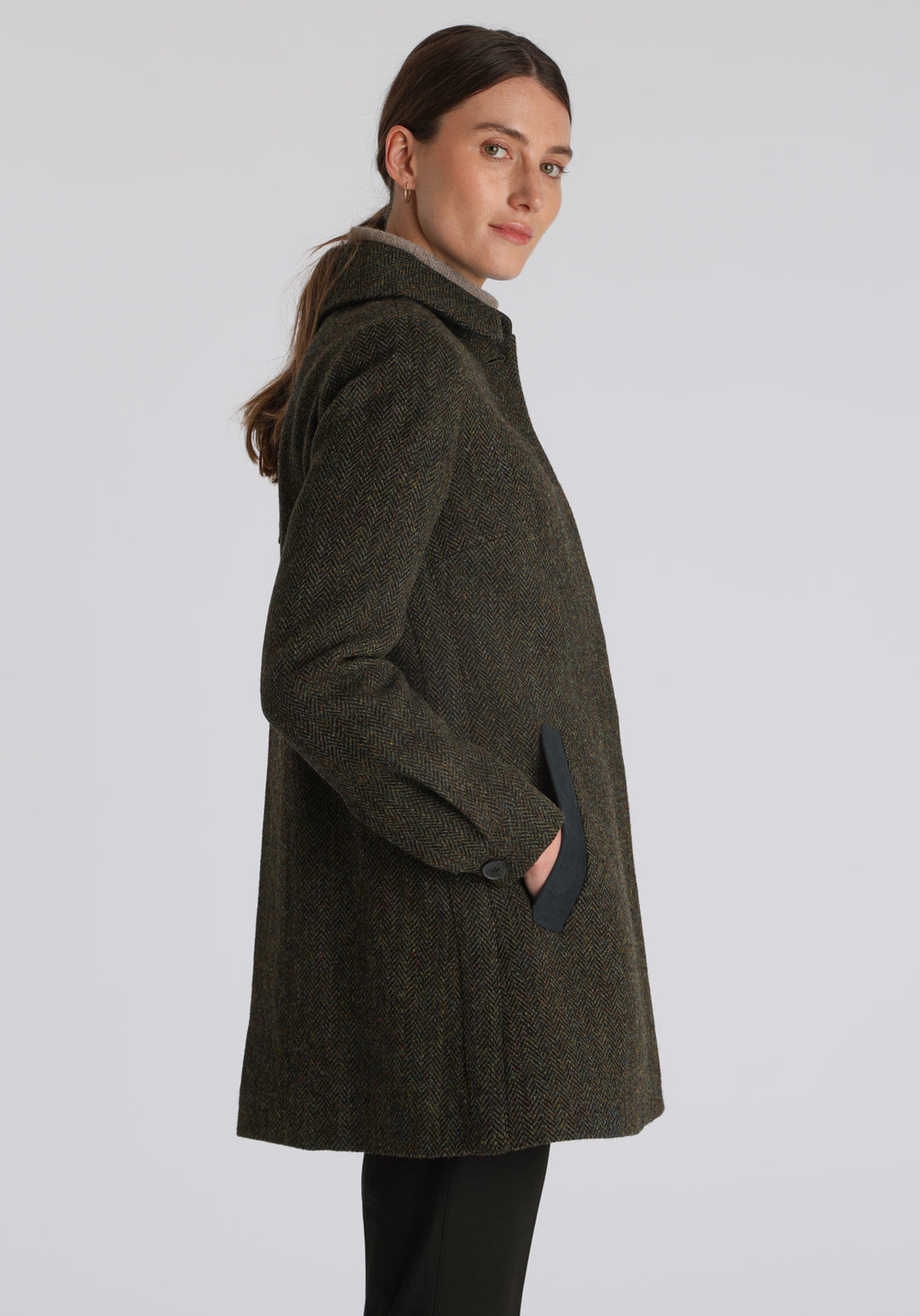 Model wearing the Cape coat in green spruce herringbone Harris Tweed, a short, structured style with a generous collar, buttoned cuffs, and a deep back pleat that creates a subtle cape-like shape.
