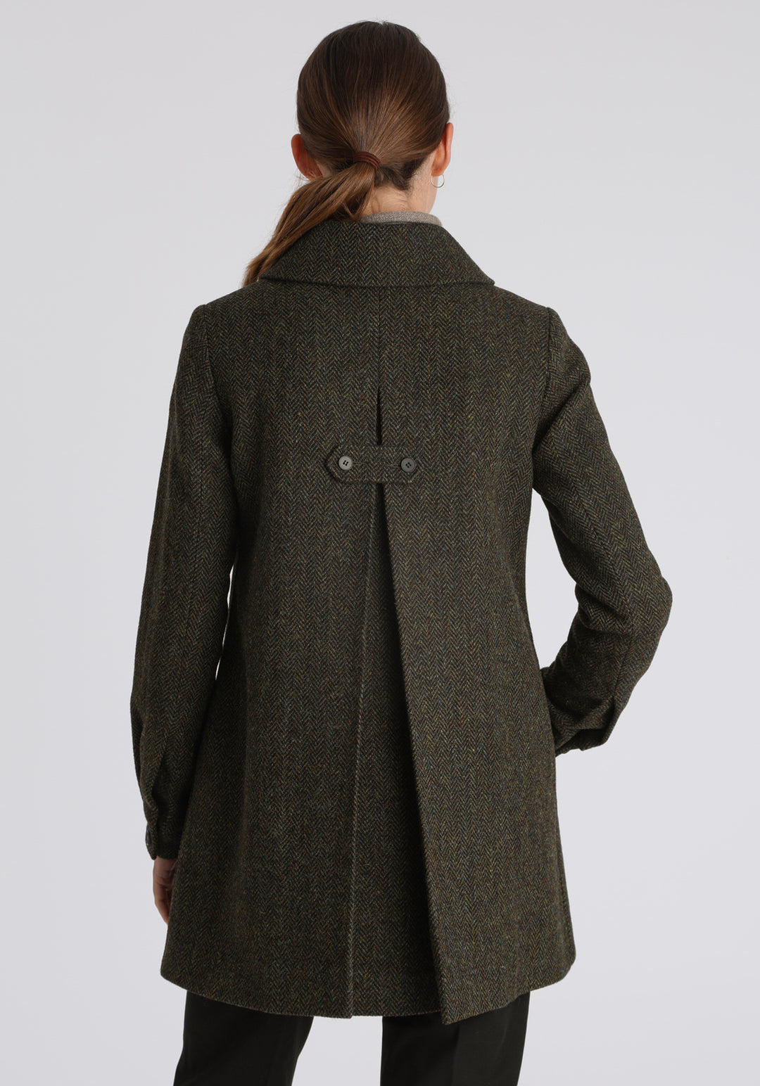 Model wearing the Cape coat in green spruce herringbone Harris Tweed, a short, structured style with a generous collar, buttoned cuffs, and a deep back pleat that creates a subtle cape-like shape.