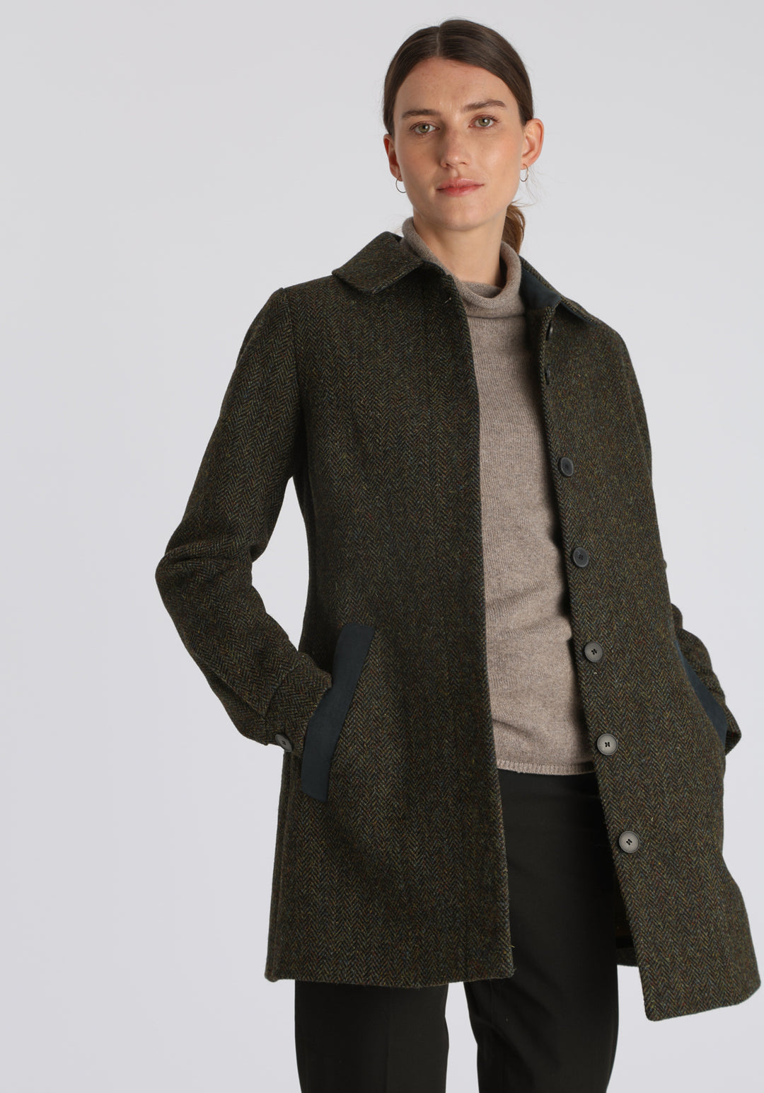 Model wearing the Cape coat in green spruce herringbone Harris Tweed, a short, structured style with a generous collar, buttoned cuffs, and a deep back pleat that creates a subtle cape-like shape.
