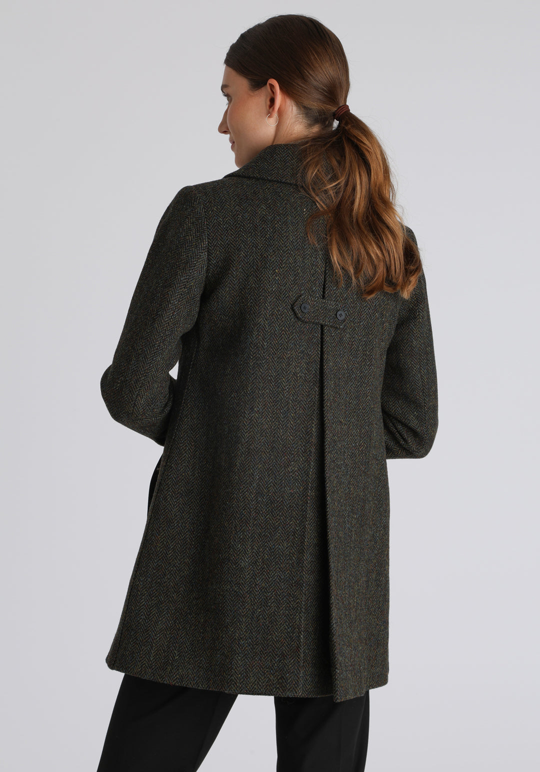 Model wearing the Cape coat in green spruce herringbone Harris Tweed, a short, structured style with a generous collar, buttoned cuffs, and a deep back pleat that creates a subtle cape-like shape.