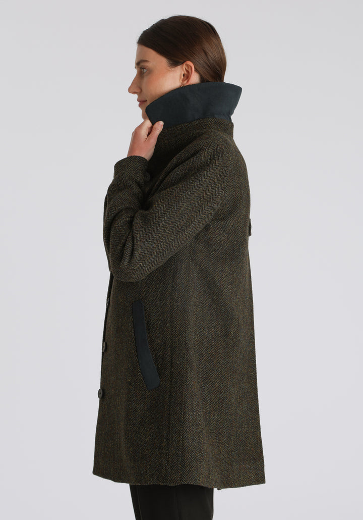 Model wearing the Cape coat in green spruce herringbone Harris Tweed, a short, structured style with a generous collar, buttoned cuffs, and a deep back pleat that creates a subtle cape-like shape.