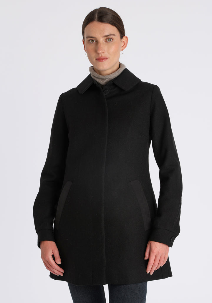 Model wearing the Cape coat in black textured merino wool, a short, structured style with a generous collar, buttoned cuffs, and a deep back pleat that creates a subtle cape-like shape.
