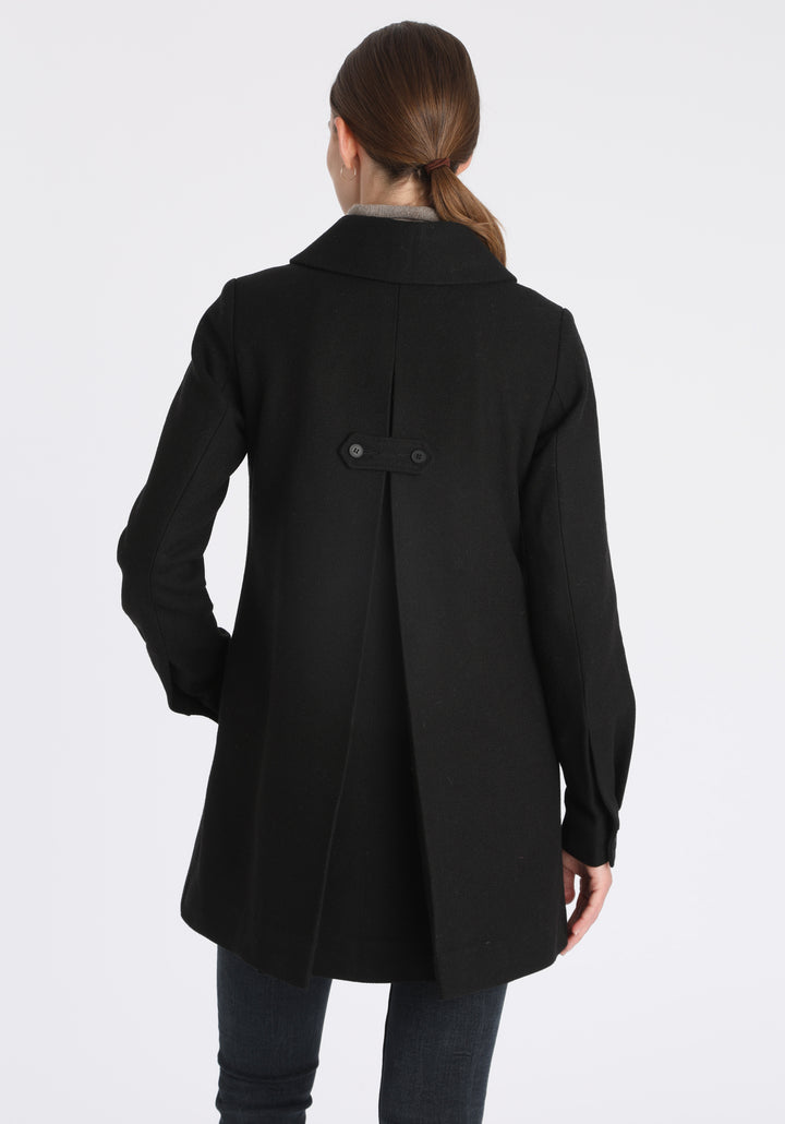 Model wearing the Cape coat in black textured merino wool, a short, structured style with a generous collar, buttoned cuffs, and a deep back pleat that creates a subtle cape-like shape.