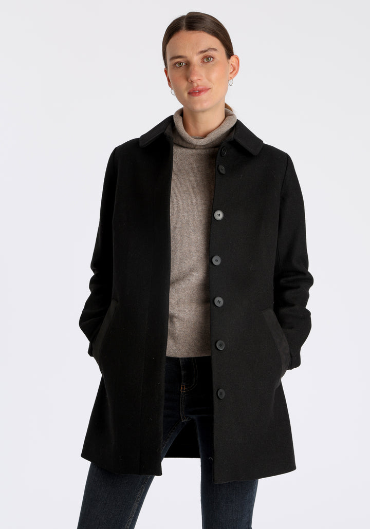 Model wearing the Cape coat in black textured merino wool, a short, structured style with a generous collar, buttoned cuffs, and a deep back pleat that creates a subtle cape-like shape.