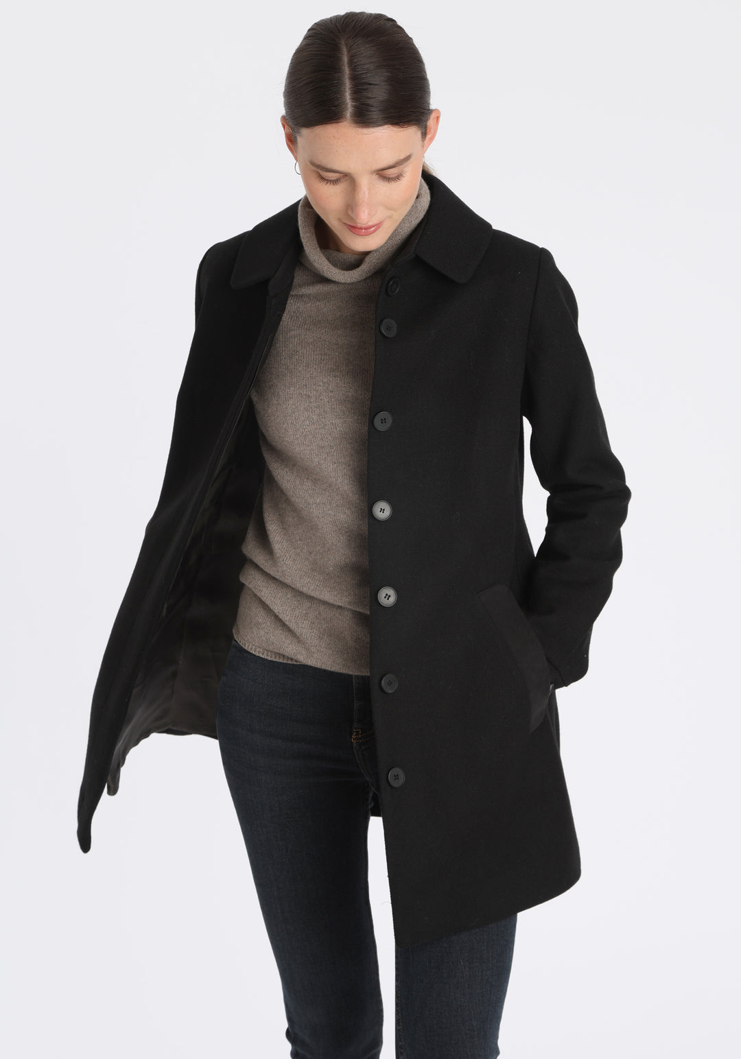 Model wearing the Cape coat in black textured merino wool, a short, structured style with a generous collar, buttoned cuffs, and a deep back pleat that creates a subtle cape-like shape.