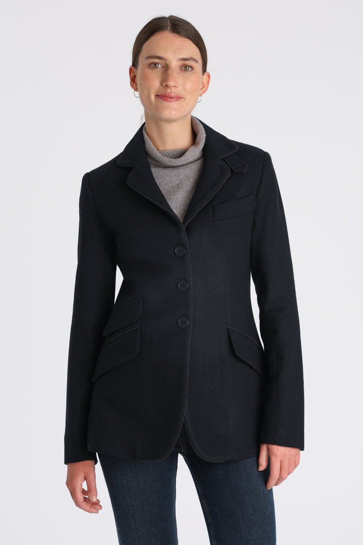 Model wearing the Cass jacket in blue spruce herringbone Harris Tweed with suede trim, a longline, relaxed boyfriend-style jacket with welt pockets, and buttoned collar tab inspired by equestrian tailoring.