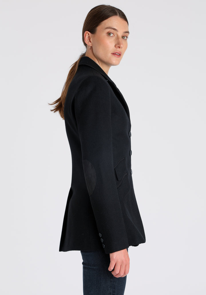 Model wearing the Cass jacket in blue spruce herringbone Harris Tweed with suede trim, a longline, relaxed boyfriend-style jacket with welt pockets, and buttoned collar tab inspired by equestrian tailoring.