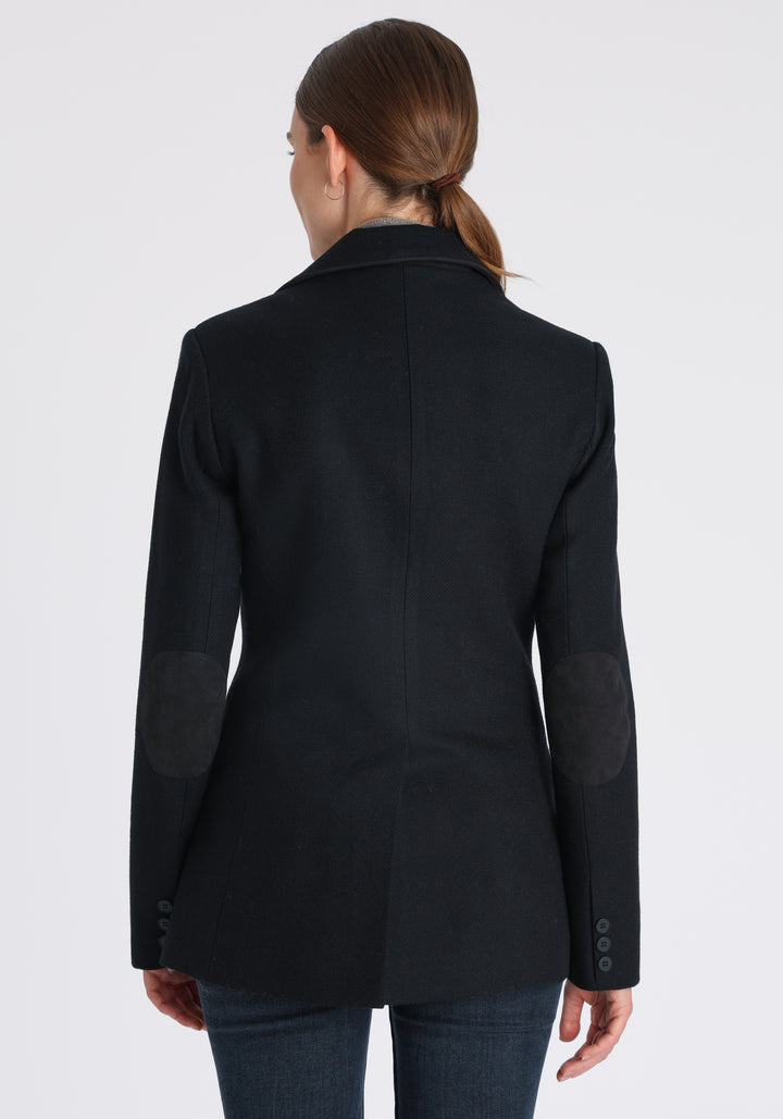 Model wearing the Cass jacket in blue spruce herringbone Harris Tweed with suede trim, a longline, relaxed boyfriend-style jacket with welt pockets, and buttoned collar tab inspired by equestrian tailoring.