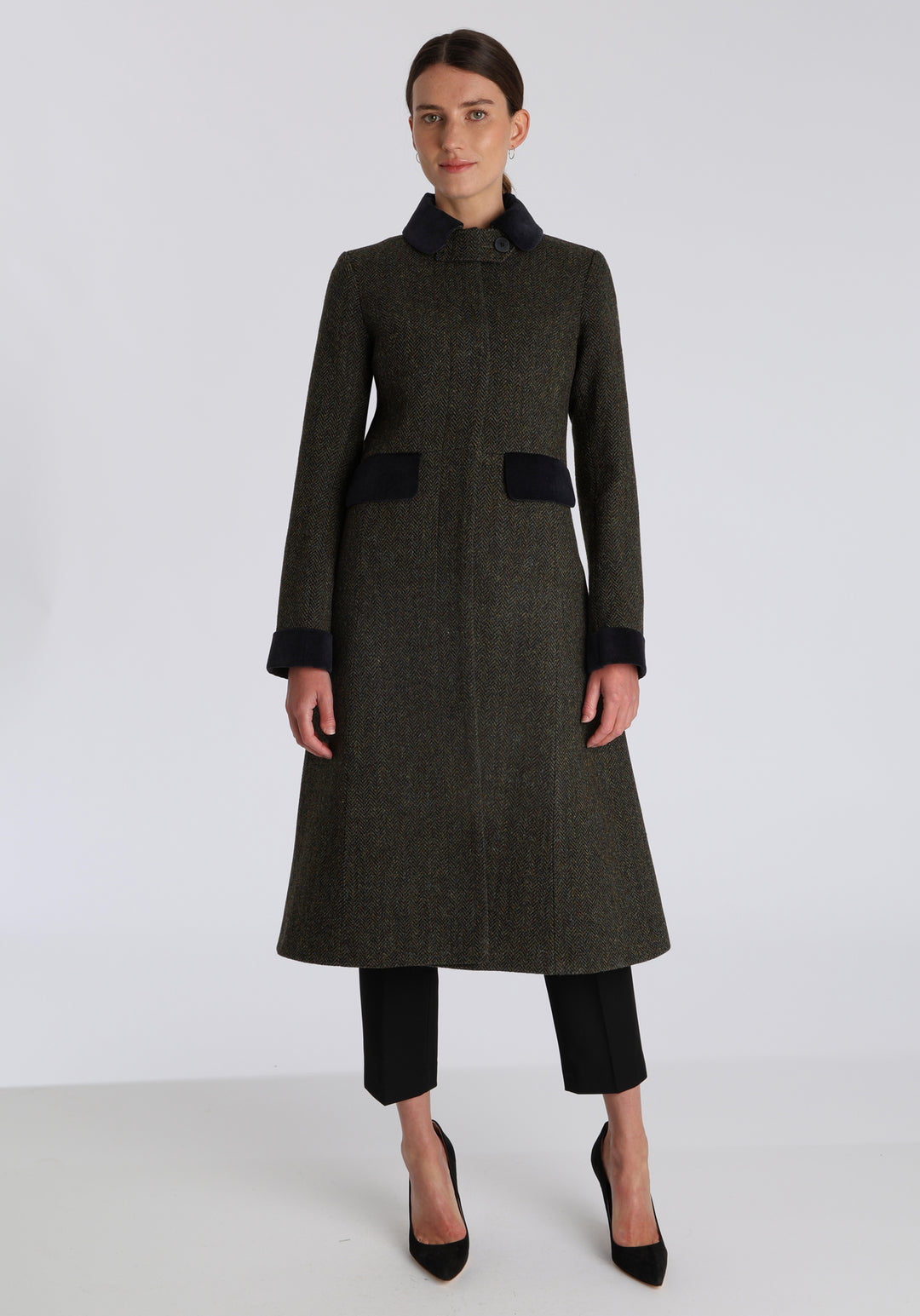Model wearing the Contrast coat in green spruce herringbone Harris Tweed with cotton velvet detailing on the collar, cuffs, back belt, and pocket flaps, a longline occasion wear style with a timeless silhouette.