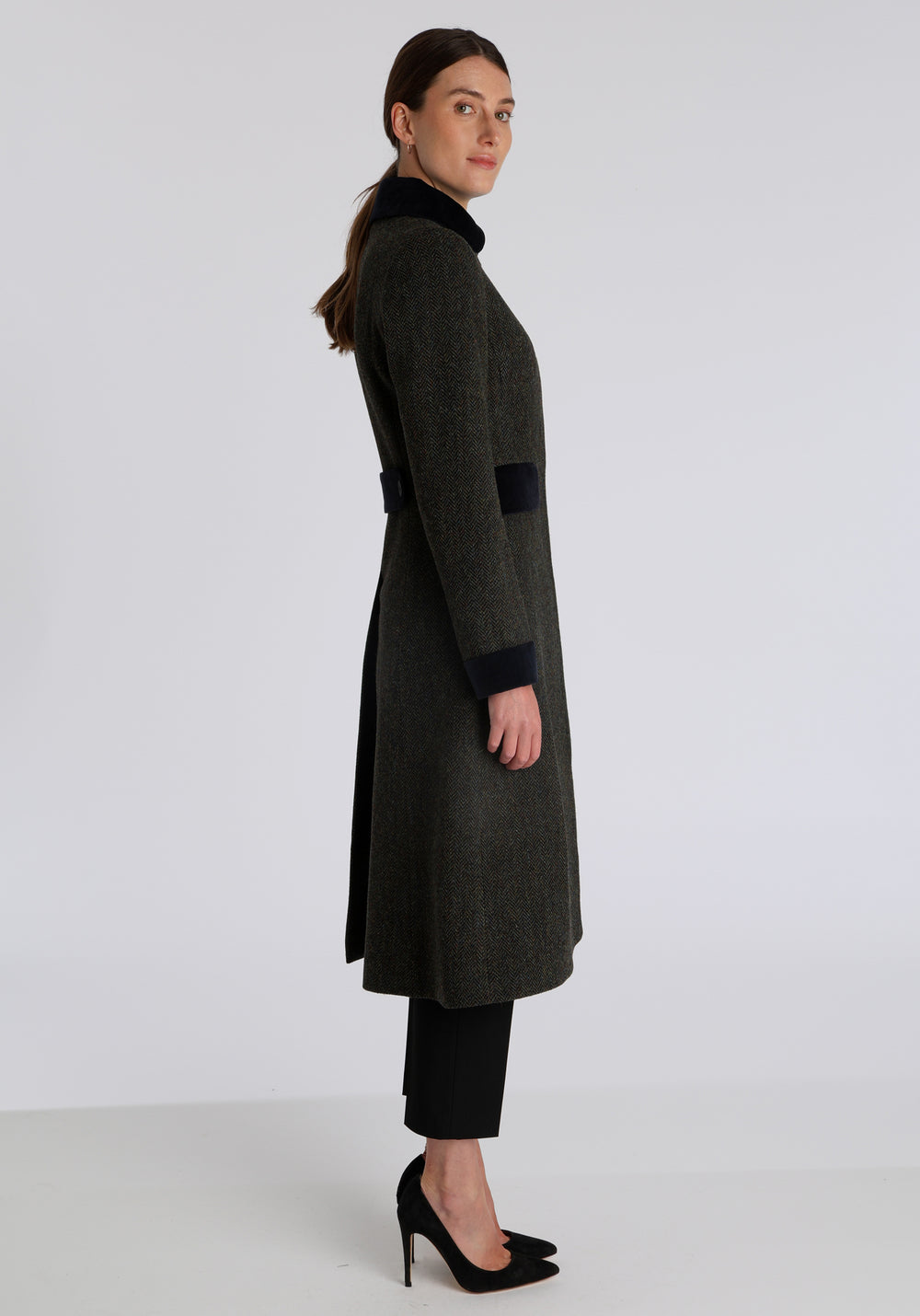 Model wearing the Contrast coat in green spruce herringbone Harris Tweed with cotton velvet detailing on the collar, cuffs, back belt, and pocket flaps, a longline occasion wear style with a timeless silhouette.
