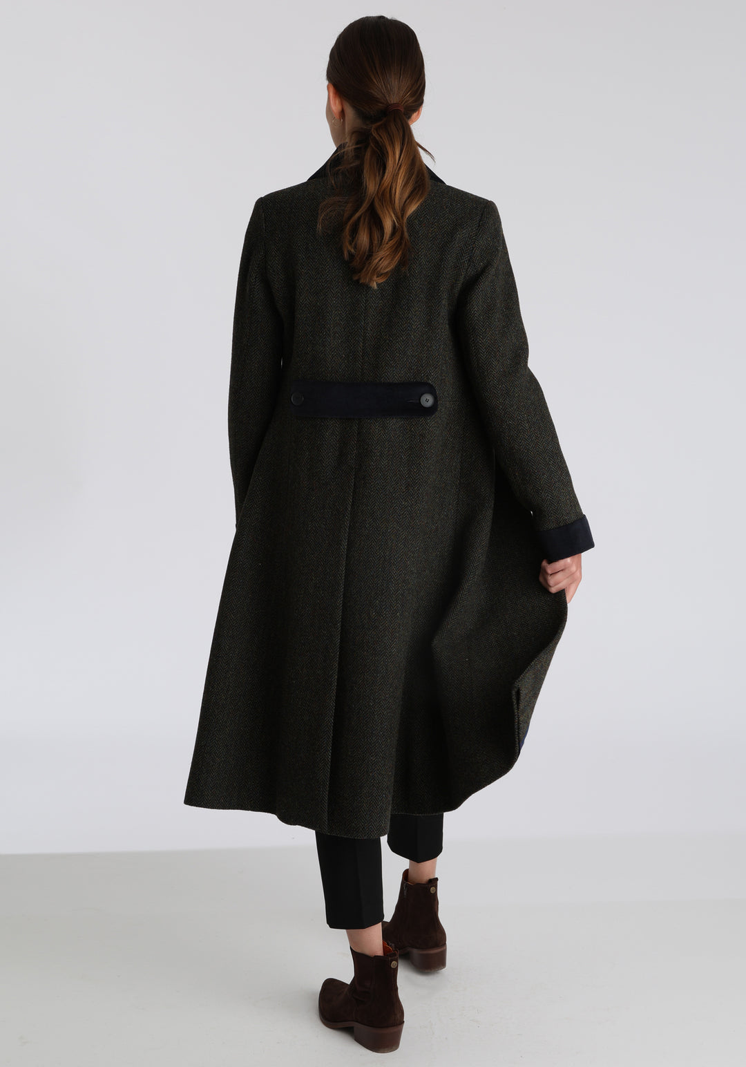 Model wearing the Contrast coat in green spruce herringbone Harris Tweed with cotton velvet detailing on the collar, cuffs, back belt, and pocket flaps, a longline occasion wear style with a timeless silhouette.