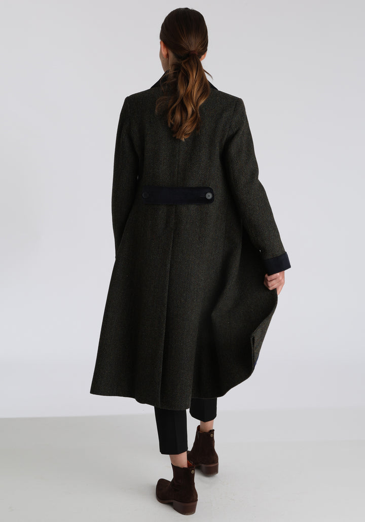 Model wearing the Contrast coat in green spruce herringbone Harris Tweed with cotton velvet detailing on the collar, cuffs, back belt, and pocket flaps, a longline occasion wear style with a timeless silhouette.