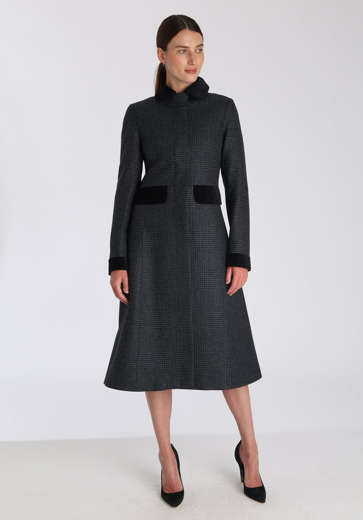 Model wearing the Contrast coat in black and charcoal grey Prince of Wales check merino wool with cotton velvet detailing on the collar, cuffs, back belt, and pocket flaps, a longline occasionwear style with a timeless silhouette.