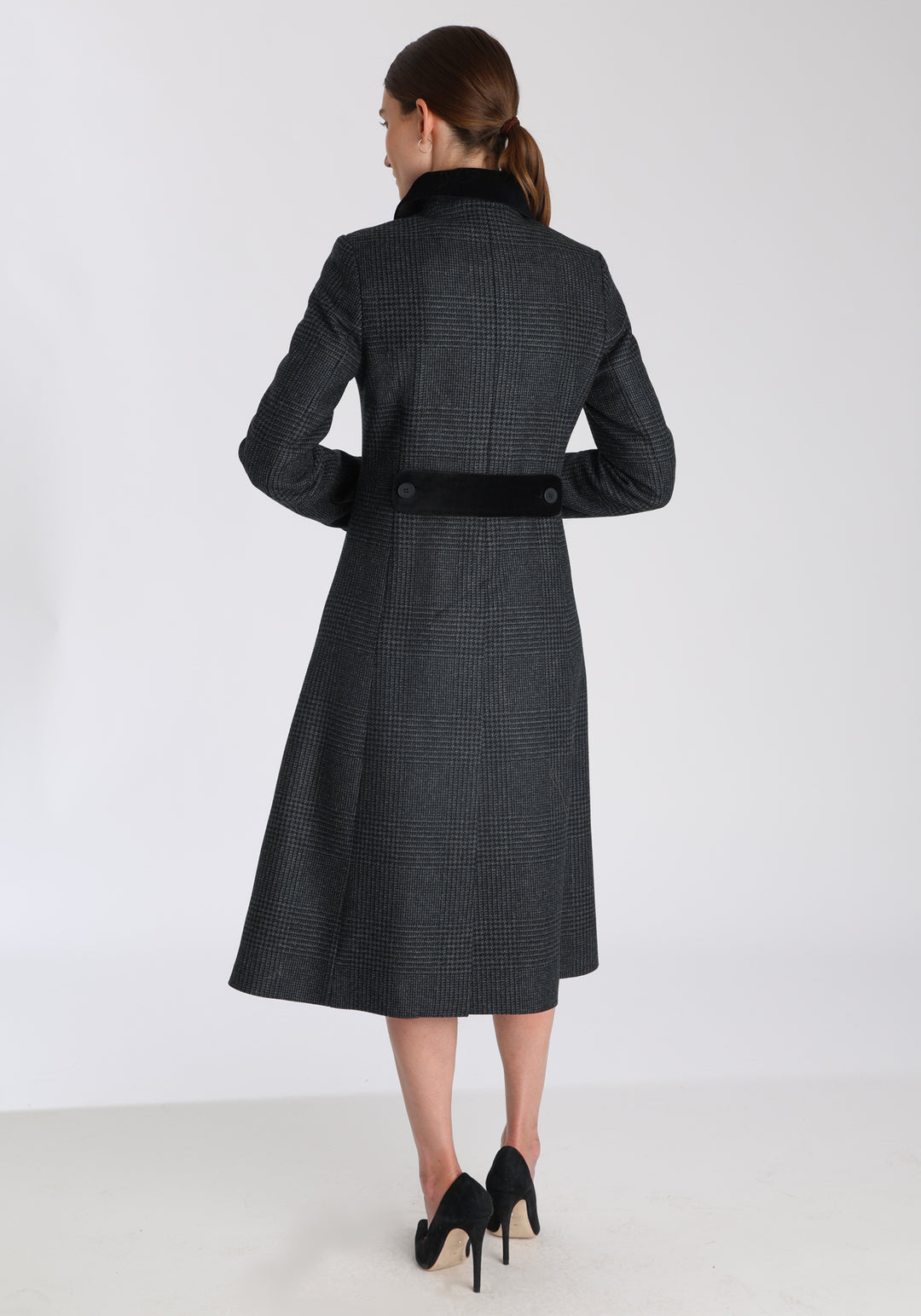 Model wearing the Contrast coat in black and charcoal grey Prince of Wales check merino wool with cotton velvet detailing on the collar, cuffs, back belt, and pocket flaps, a longline occasionwear style with a timeless silhouette.