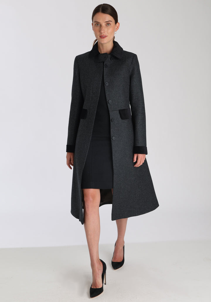 Model wearing the Contrast coat in black and charcoal grey Prince of Wales check merino wool with cotton velvet detailing on the collar, cuffs, back belt, and pocket flaps, a longline occasionwear style with a timeless silhouette.