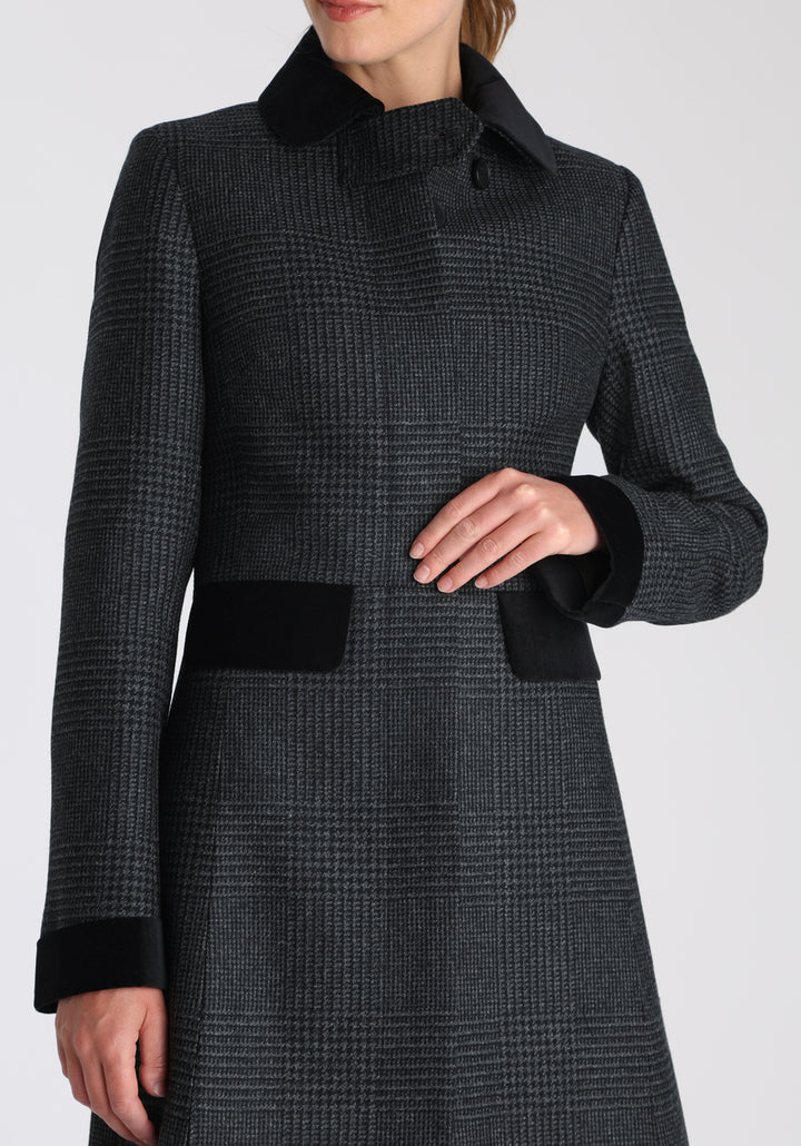 Model wearing the Contrast coat in black and charcoal grey Prince of Wales check merino wool with cotton velvet detailing on the collar, cuffs, back belt, and pocket flaps, a longline occasionwear style with a timeless silhouette.