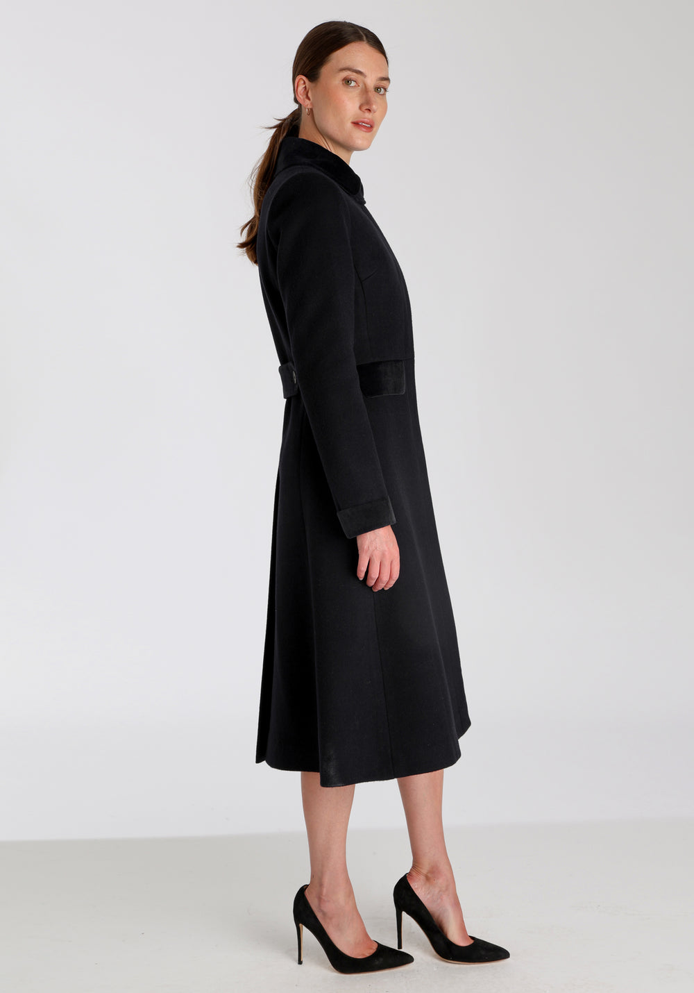 Model wearing the Contrast coat in black wool cashmere with cotton velvet detailing on the collar, cuffs, back belt, and pocket flaps, a longline occasionwear style with a timeless silhouette.