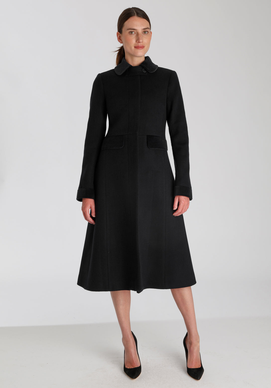 Model wearing the Contrast coat in black wool cashmere with cotton velvet detailing on the collar, cuffs, back belt, and pocket flaps, a longline occasionwear style with a timeless silhouette.