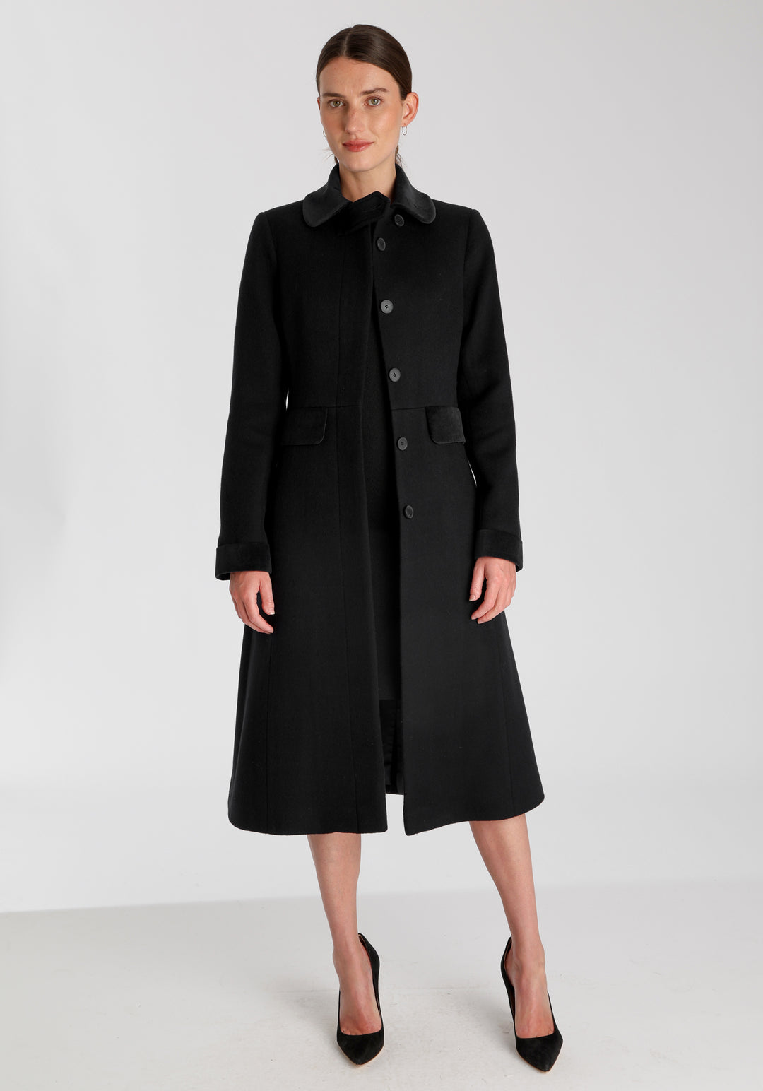 Model wearing the Contrast coat in black wool cashmere with cotton velvet detailing on the collar, cuffs, back belt, and pocket flaps, a longline occasionwear style with a timeless silhouette.
