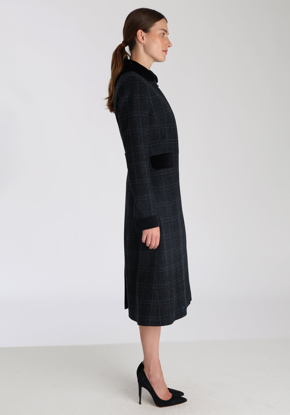 Model wearing the Contrast coat in black with faint overcheck, a long tailored style with black cotton velvet contrasting pockets, cuffs and back belt with a deep back vent for ease of movement.