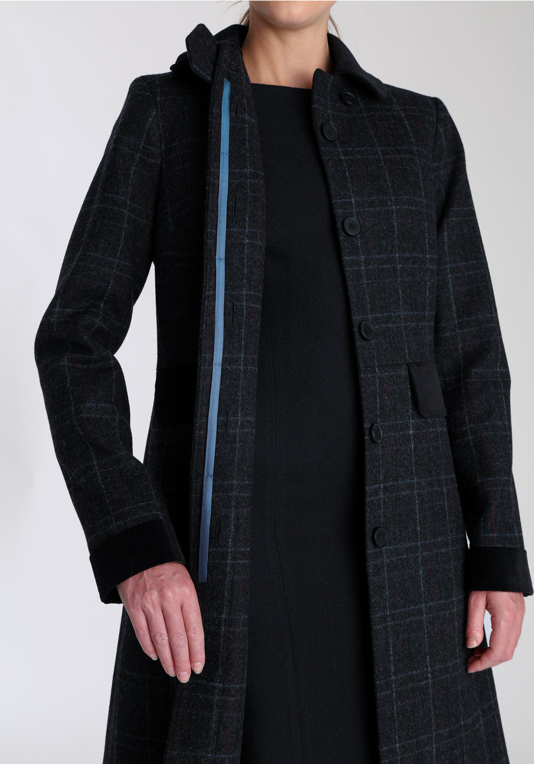 Model wearing the Contrast coat in black with faint overcheck, a long tailored style with black cotton velvet contrasting pockets, cuffs and back belt with a deep back vent for ease of movement.