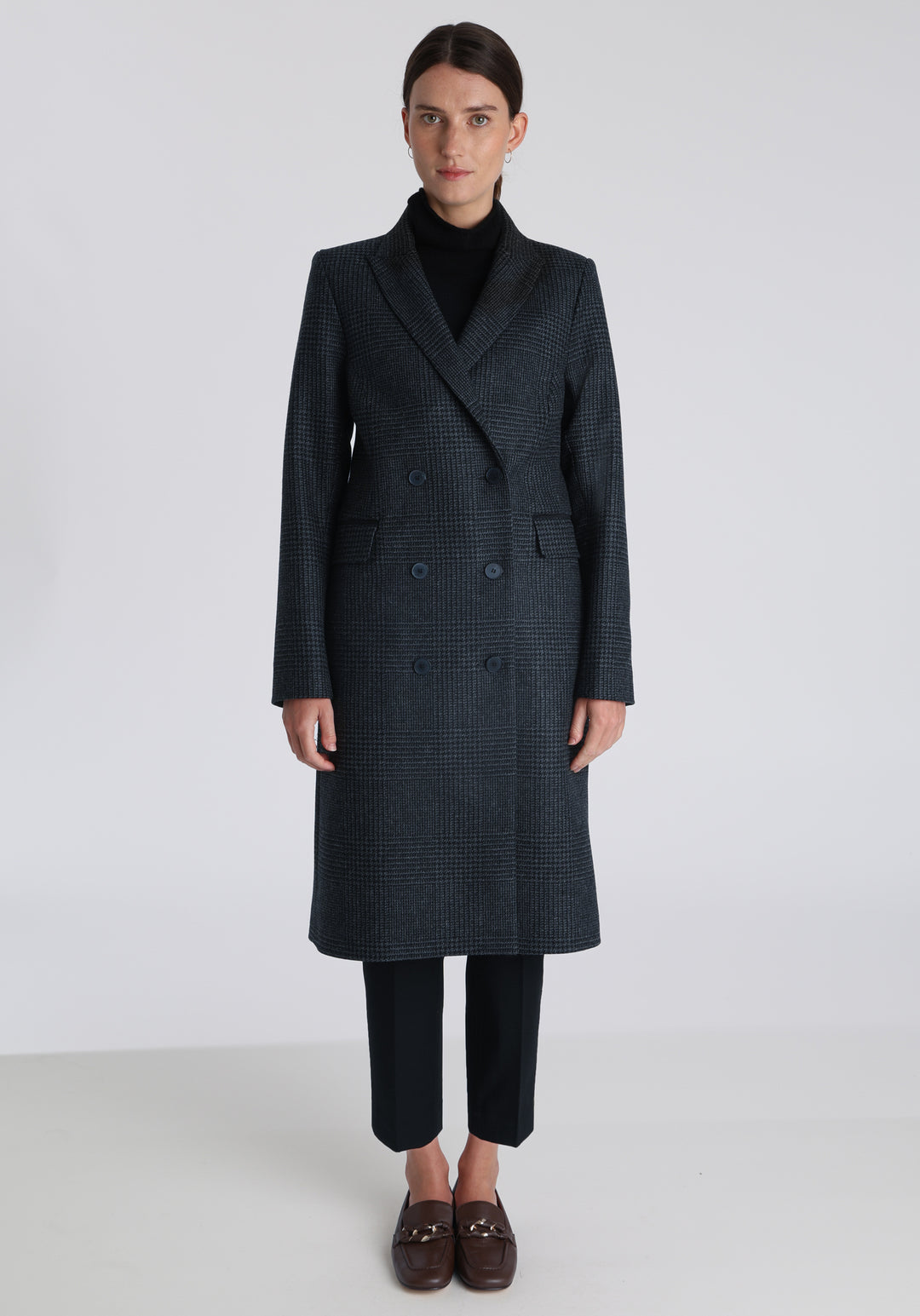 Model wearing the Cully coat in black and charcoal grey Prince of Wales check wool, a knee-length, double-breasted style with pointed lapels and a structured, straight-cut silhouette designed to flatter.