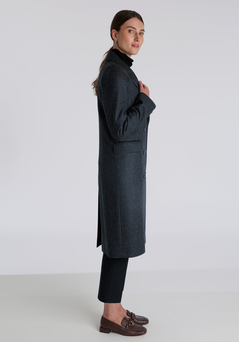 Model wearing the Cully coat in black and charcoal grey Prince of Wales check wool, a knee-length, double-breasted style with pointed lapels and a structured, straight-cut silhouette designed to flatter.