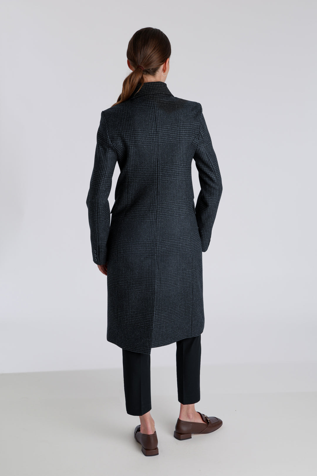 Model wearing the Cully coat in black and charcoal grey Prince of Wales check wool, a knee-length, double-breasted style with pointed lapels and a structured, straight-cut silhouette designed to flatter.