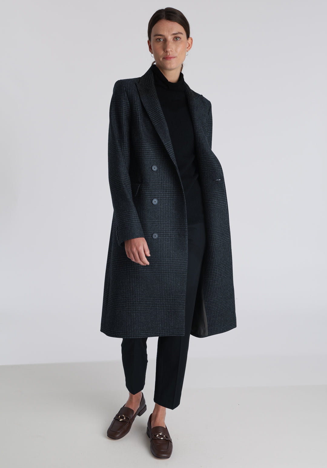 Model wearing the Cully coat in black and charcoal grey Prince of Wales check wool, a knee-length, double-breasted style with pointed lapels and a structured, straight-cut silhouette designed to flatter.