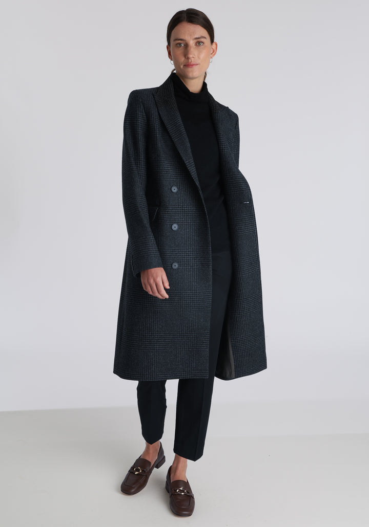 Model wearing the Cully coat in black and charcoal grey Prince of Wales check wool, a knee-length, double-breasted style with pointed lapels and a structured, straight-cut silhouette designed to flatter.